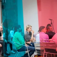Through a curtain of blue, red and yellow plastic strips you can see a group of people sitting on metal chairs, listening attentively.
