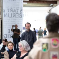 Attracting the attention of the audience, artist and activist Tanja Krone speaks to them. She stands with her back to the camera, with numerous city coats of arms attached to her coat.