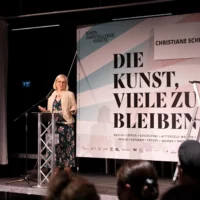Dr. Christiane Schenderlein, member of the Bundestag and cultural policy spokesperson for the CDU, stands in front of a billboard with the inscription "DIE KUNST, VIELE ZU BLEIBEN" and gives a keynote speech to the audience while standing at a lectern.