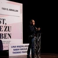 Fadi El Abdallah stands at the lectern during his presentation on stage. The billboard with the inscription "DIE KUNST, VIELE ZU BLEIBEN" and his name can be seen in the background.