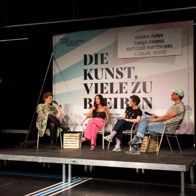 The panel guests Antonie Rietzschel, Mirna Funk, Tanja Krone and Ludwig Henne sit next to each other in a semi-circle on a stage. Behind them is a large billboard with the inscription "DIE KUNST, VIELE ZU BLEIBEN".