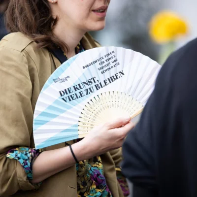 One person is holding a fan with the inscription "DIE KUNST, VIELE ZU BLEIBEN" and is fanning themselves while talking to another person.